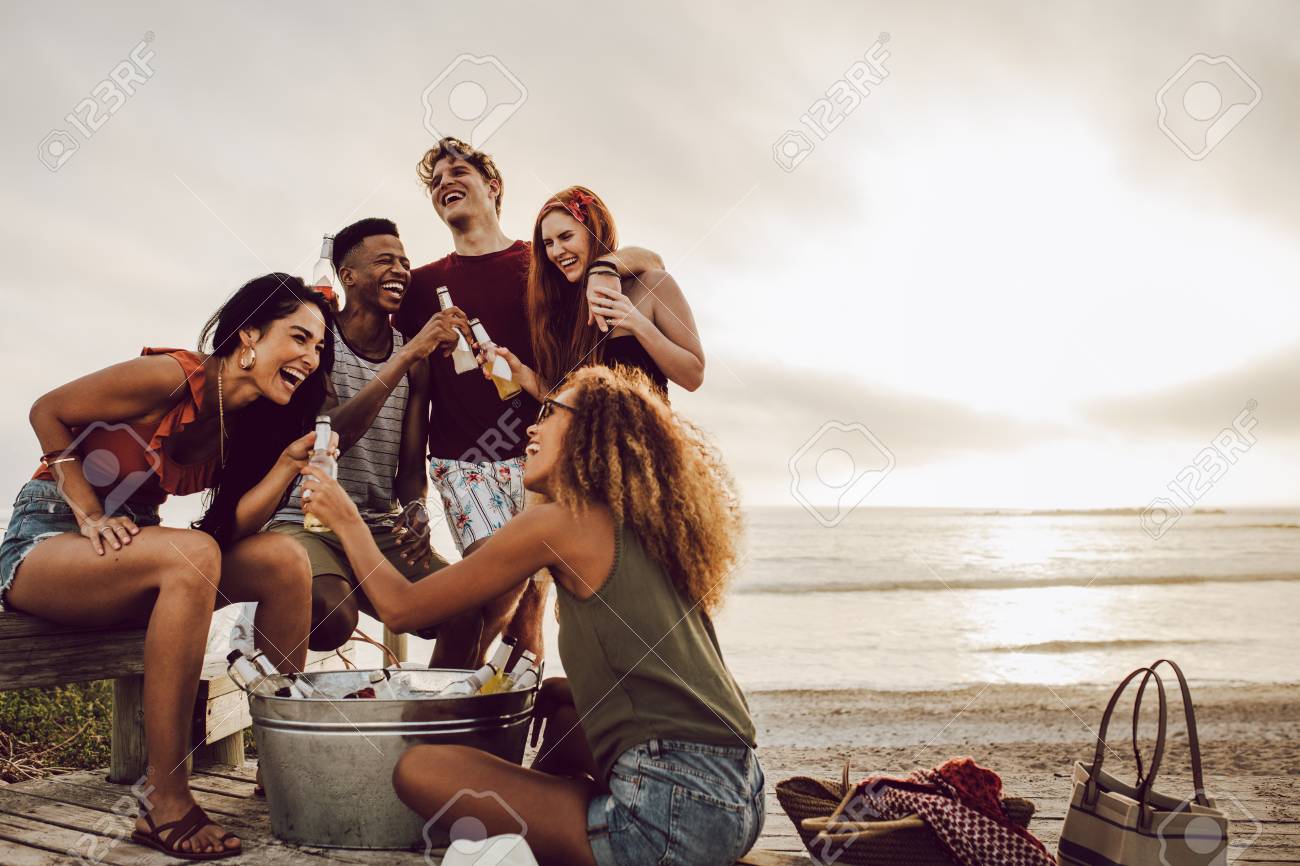 Smiling young woman with beer bottle and friends standing by on the beach. - 124972526 Smiling young woman with beer bottle and friends standing by on the beach. - 124972526
