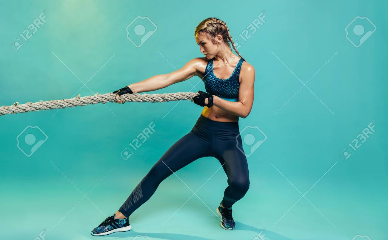 Tough young woman exercising with battling rope in studio. Healthy sports woman working out with battle rope over blue background. - 127402015 Tough young woman exercising with battling rope in studio. Healthy sports woman working out with battle rope over blue background. - 127402015