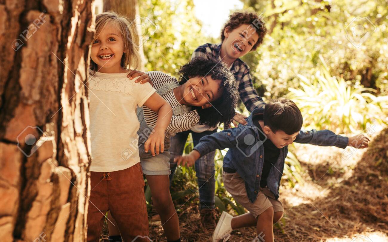 Cute smiling kids peeking out from behind the tree in the park. Group of children enjoying playing hide and seek in a forest. - 127853839 Cute smiling kids peeking out from behind the tree in the park. Group of children enjoying playing hide and seek in a forest. - 127853839