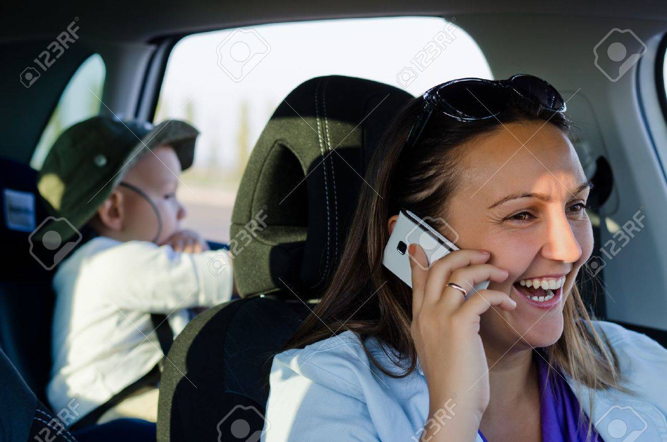 Woman driver with a young boy strapped into a child safety seat in the back of the car laughing and chatting on her mobile phone - 17657775 Woman driver with a young boy strapped into a child safety seat in the back of the car laughing and chatting on her mobile phone - 17657775