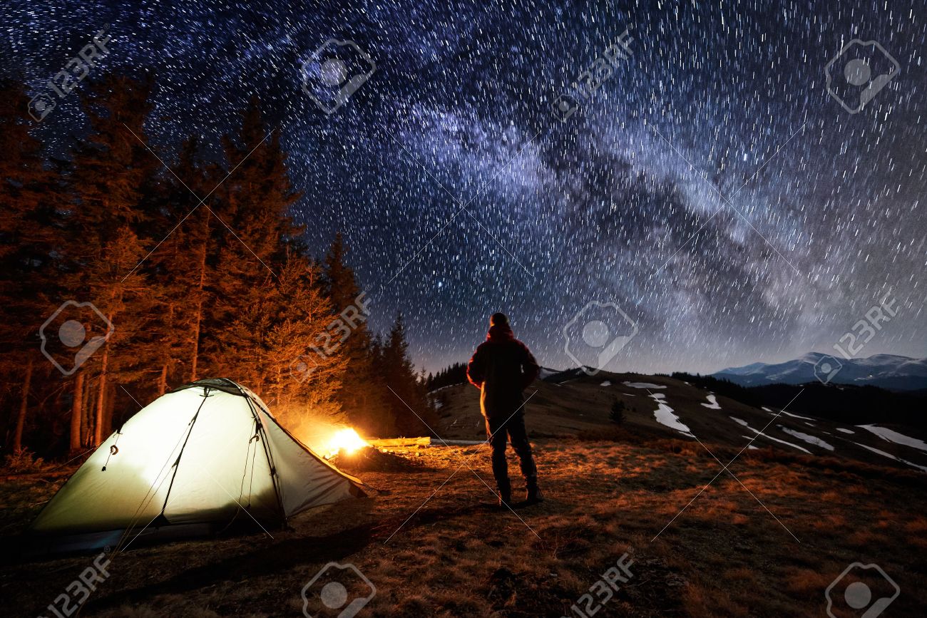 Male tourist have a rest in his camp near the forest at night. Man standing near campfire and tent under beautiful night sky full of stars and milky way, and enjoying night scene. Long exposure - 80507834 Male tourist have a rest in his camp near the forest at night. Man standing near campfire and tent under beautiful night sky full of stars and milky way, and enjoying night scene. Long exposure - 80507834