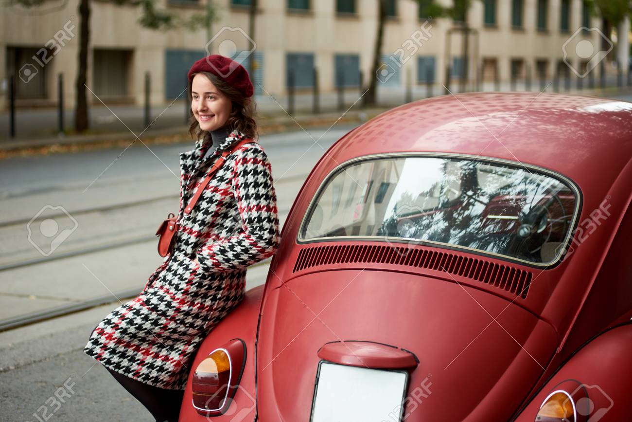 後ろの赤いレトロな車のクローズアップと その近くにかわいい女の子 女の子はコートを着て 赤い色調のベレーを着ている ヴィンテージスタイル の写真素材 画像素材 Image