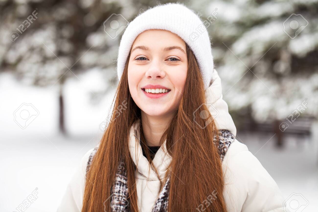 Close up portrait of a young beautiful girl in a white down jacket posing in a winter park - 138434316 Close up portrait of a young beautiful girl in a white down jacket posing in a winter park - 138434316