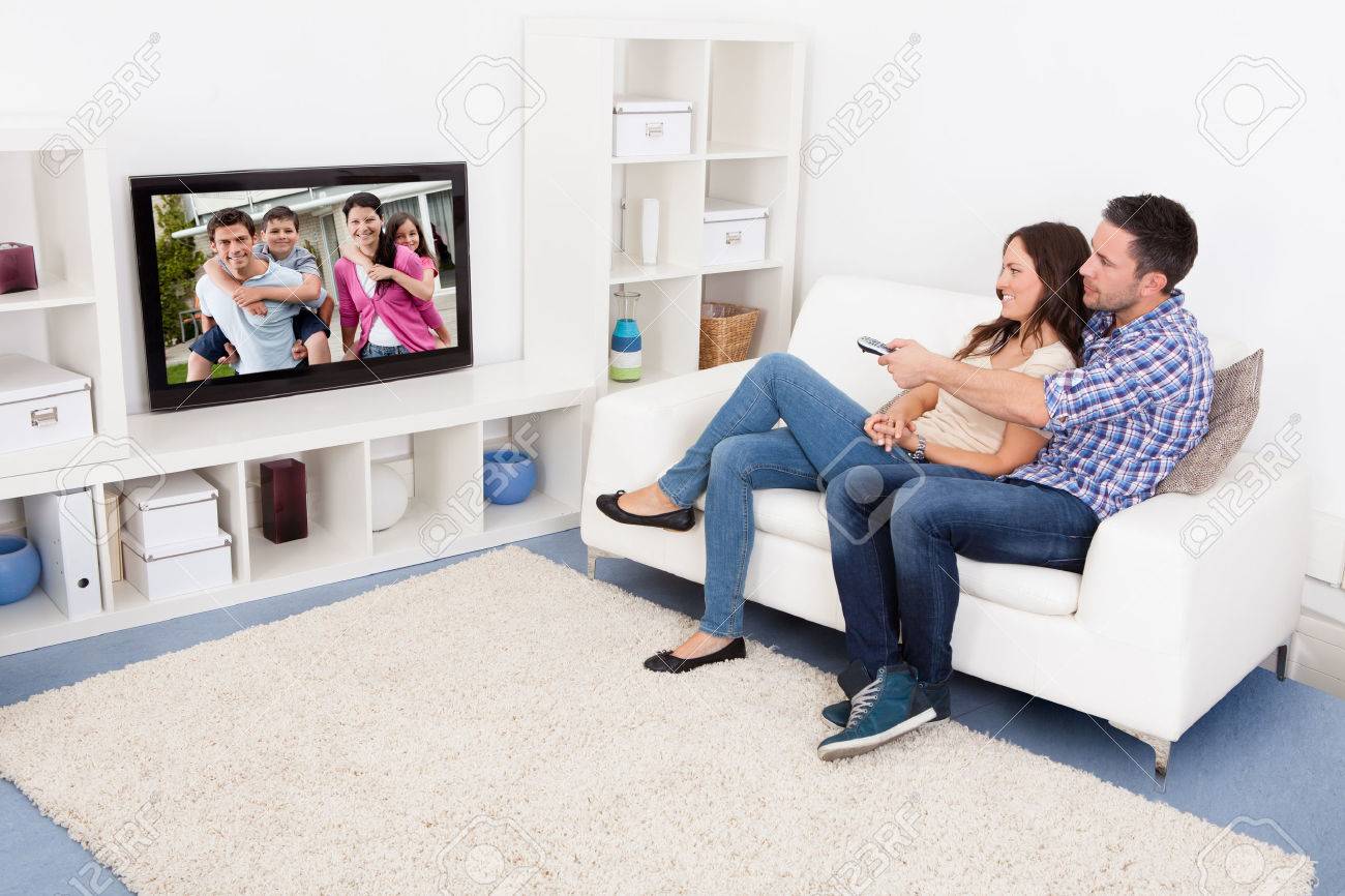 Happy Young Couple In Livingroom Sitting On Couch Watching Television - 24123100 Happy Young Couple In Livingroom Sitting On Couch Watching Television - 24123100