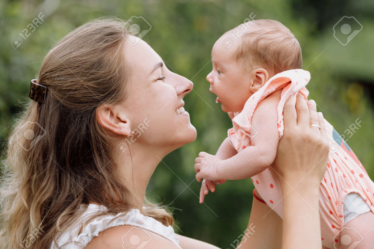 Mother is holding and kissing her baby girl outdoors on summer day. Summer family time and fresh air concept. - 156060285 Mother is holding and kissing her baby girl outdoors on summer day. Summer family time and fresh air concept. - 156060285