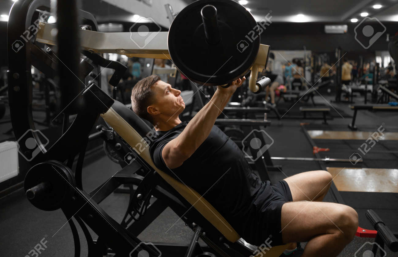 A young male athlete on the simulator in a sitting position shakes the muscles of his arms and chest. Athlete side view. - 165701552 A young male athlete on the simulator in a sitting position shakes the muscles of his arms and chest. Athlete side view. - 165701552