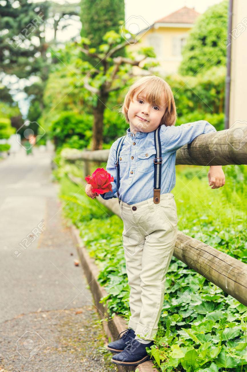 かわいい少年のサスペンダーのズボンを身に着けている赤いバラ ヴィンテージのレトロなスタイルを保持している屋外のポートレート の写真素材 画像素材 Image