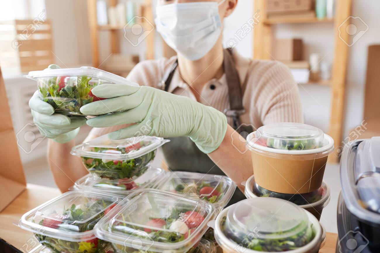 Close-up of young woman in protective mask and gloves packing fresh vegetables in boxes while sitting at the table - 150090558 Close-up of young woman in protective mask and gloves packing fresh vegetables in boxes while sitting at the table - 150090558