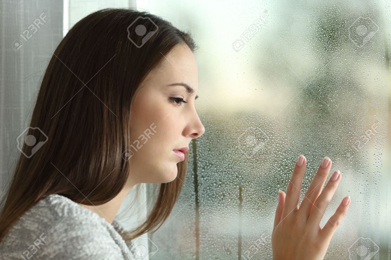 Sad woman looking the rain falling through a window at home or hotel - 52549293 Sad woman looking the rain falling through a window at home or hotel - 52549293