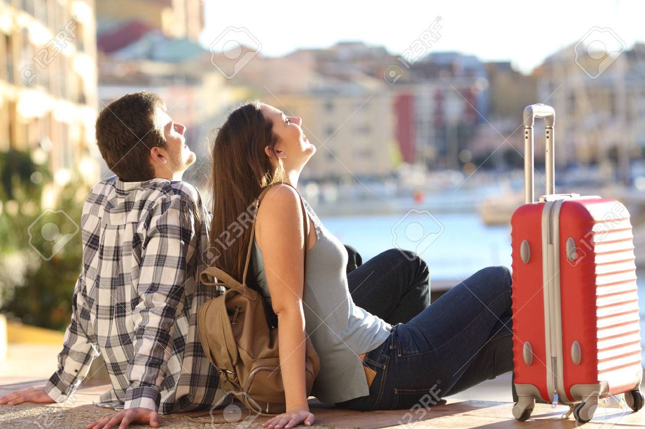 Side view of a couple of 2 tourists with a suitcase sitting relaxing and enjoying vacations in a colorful promenade. Tourism concept - 56102111 Side view of a couple of 2 tourists with a suitcase sitting relaxing and enjoying vacations in a colorful promenade. Tourism concept - 56102111