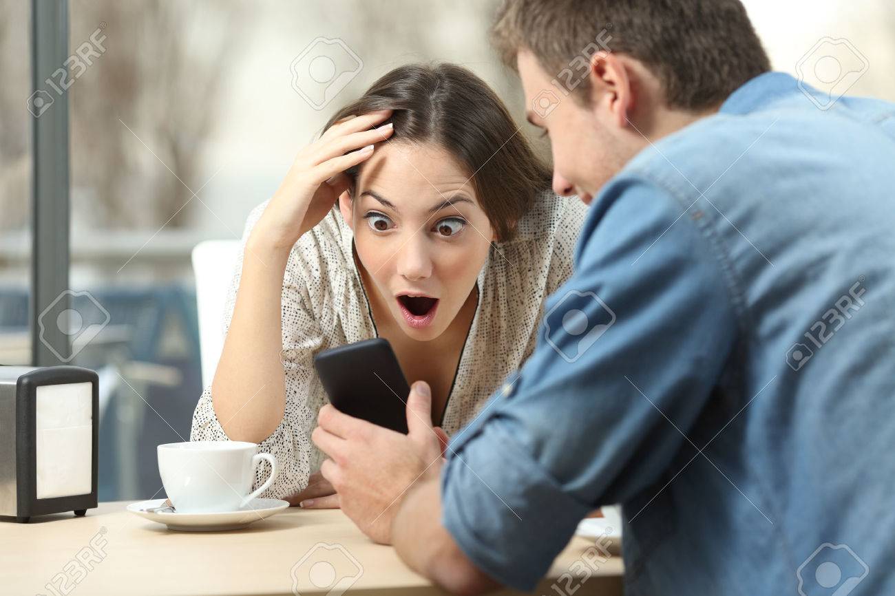 Surprised couple meeting in a coffee shop watching media content in a smart phone - 56102116 Surprised couple meeting in a coffee shop watching media content in a smart phone - 56102116