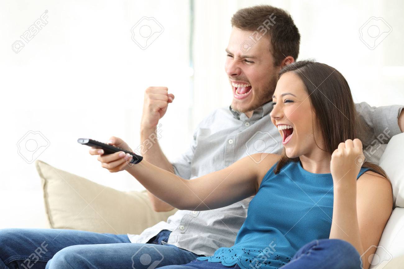 Excited couple watching tv together sitting on a sofa in the living room at home - 68711020 Excited couple watching tv together sitting on a sofa in the living room at home - 68711020
