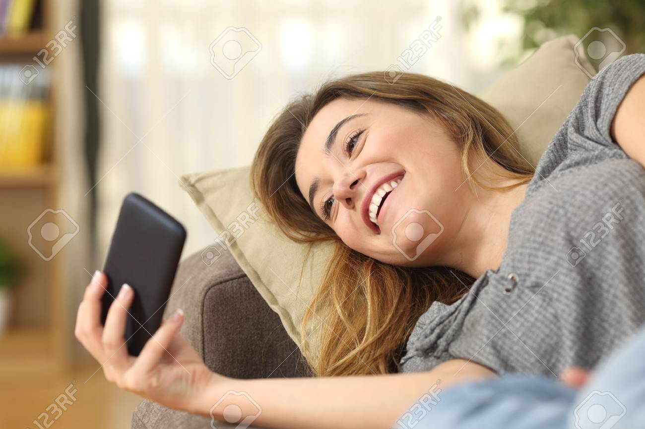 Happy Teen Watching Videos On Line In A Smart Phone Lying On A Couch In The Happy Teen Watching Videos On Line In A Smart Phone Lying On A Couch In The