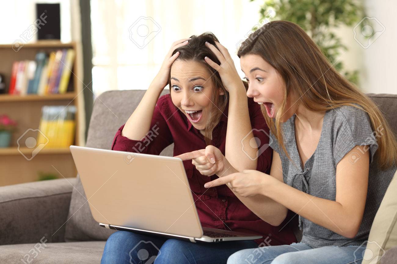 Two astonished roommates watching media content on line with a laptop sitting on a couch in the living room in a house interior - 71234177 Two astonished roommates watching media content on line with a laptop sitting on a couch in the living room in a house interior - 71234177