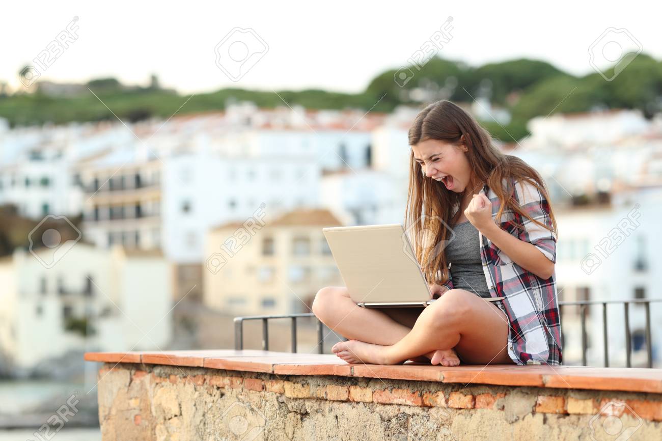 Excited teenage girl checking laptop content sitting on a ledge in a coast town on vacation - 114106183 Excited teenage girl checking laptop content sitting on a ledge in a coast town on vacation - 114106183
