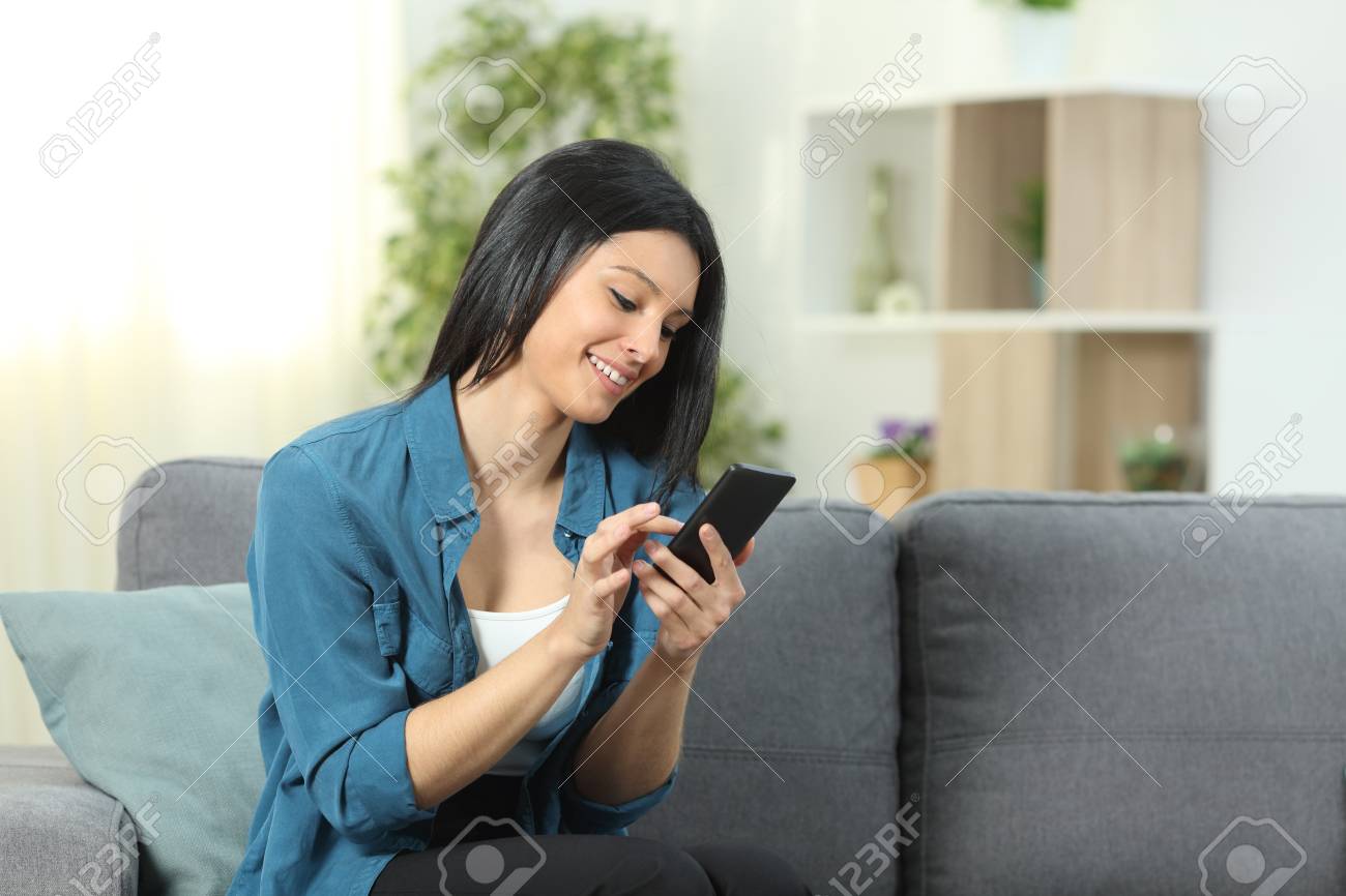 Happy woman using a smart phone sitting on a couch in the living room at home - 116123948 Happy woman using a smart phone sitting on a couch in the living room at home - 116123948