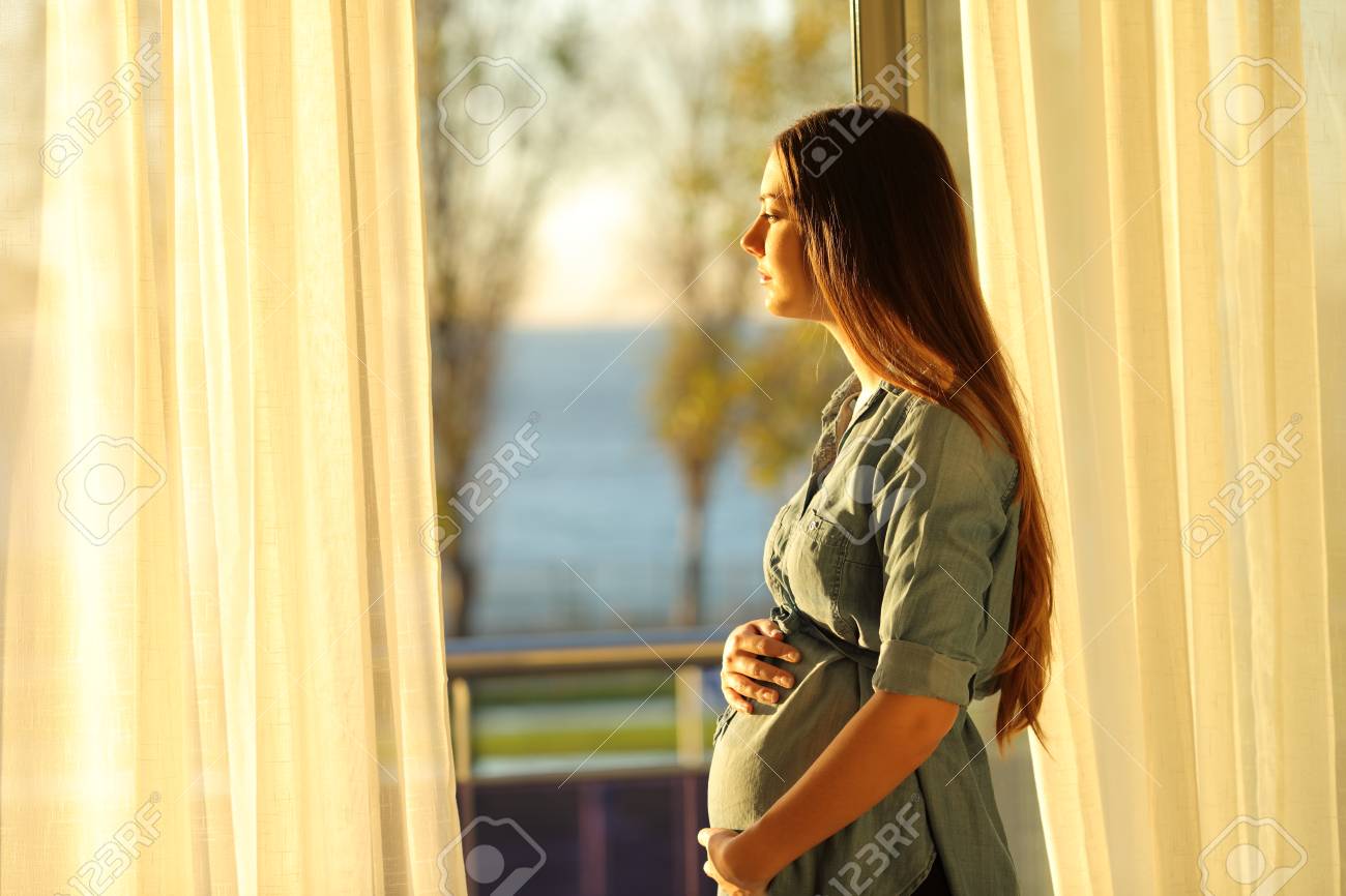 Side view portrait of a serious pregnant woman looking through a window at sunset - 117941587 Side view portrait of a serious pregnant woman looking through a window at sunset - 117941587