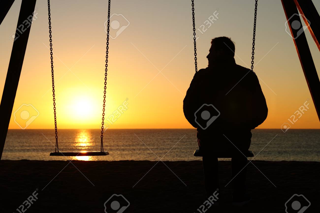 Back view backlighting silhouette of a man alone on a swing looking at empty seat at sunset on the beach in winter - 117963685 Back view backlighting silhouette of a man alone on a swing looking at empty seat at sunset on the beach in winter - 117963685