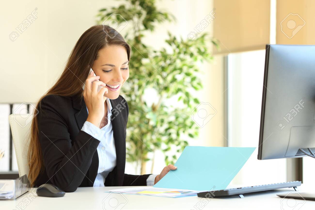 Happy office worker checking document talking on phone at workplace - 122061176 Happy office worker checking document talking on phone at workplace - 122061176