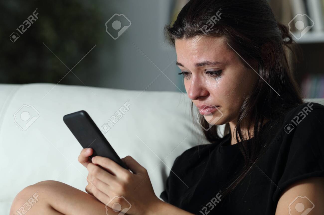 Sad teenage girl checking smart phone cyber bullying messages sitting on a couch in the dark at home - 128723134 Sad teenage girl checking smart phone cyber bullying messages sitting on a couch in the dark at home - 128723134