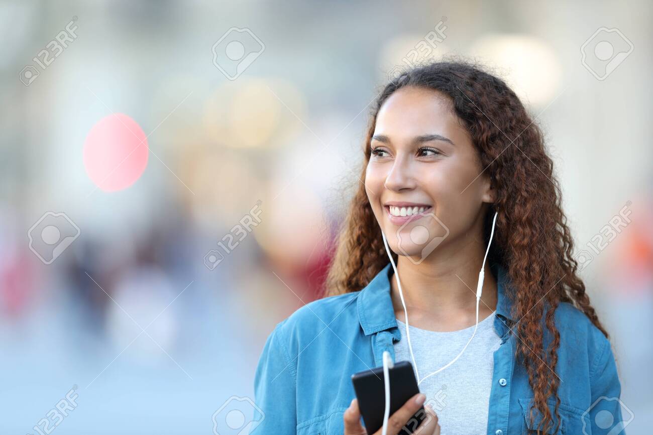 Front view portrait of a mixed race woman listening to music looking at side in the street - 129968499 Front view portrait of a mixed race woman listening to music looking at side in the street - 129968499