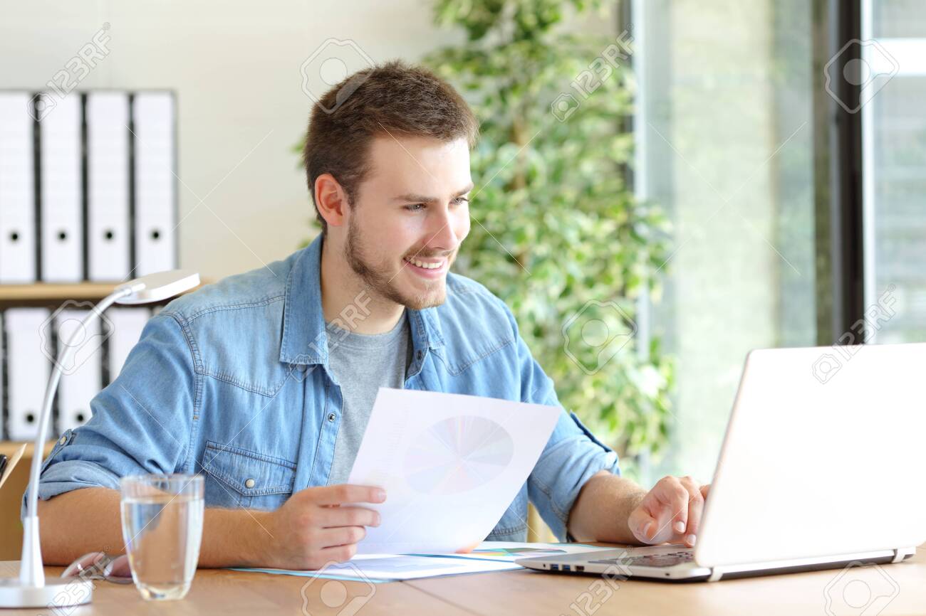 Casual happy entrepreneur working using laptop holding document comparing data at office - 133951673 Casual happy entrepreneur working using laptop holding document comparing data at office - 133951673