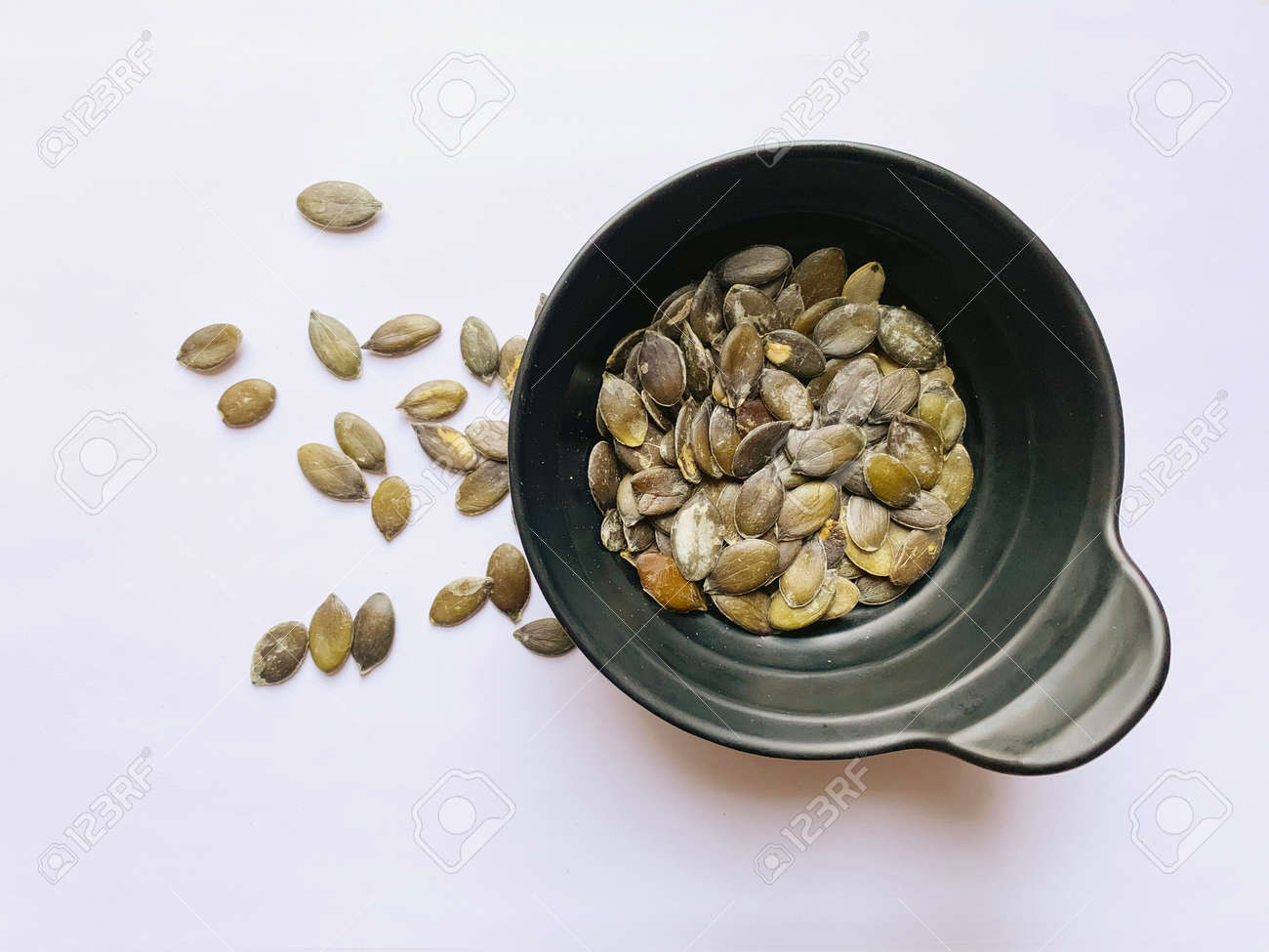 Styrian pumpkin seeds in a black cup and scattered on a white background. View from above. - 169603136 Styrian pumpkin seeds in a black cup and scattered on a white background. View from above. - 169603136
