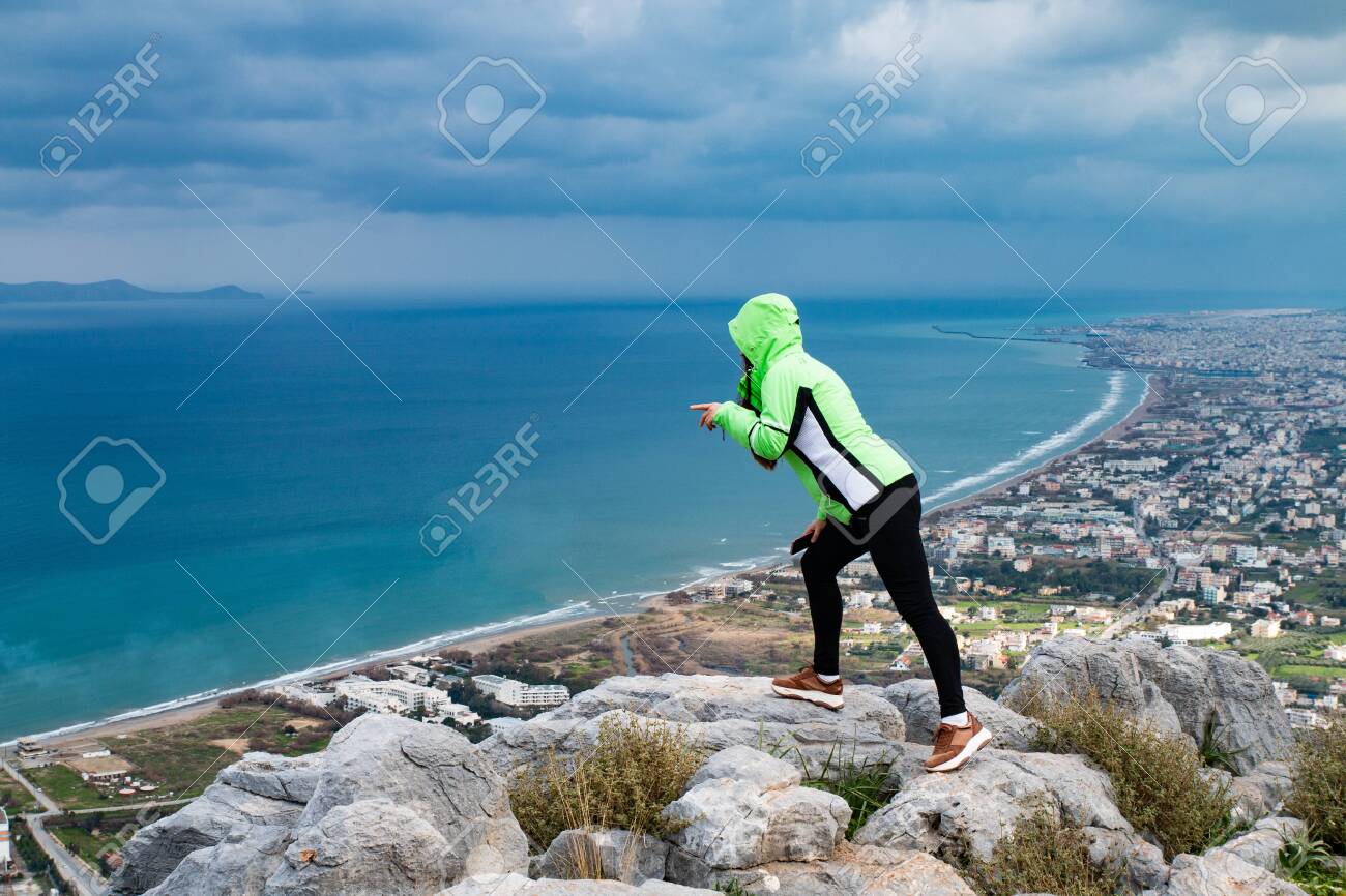 A young woman standing on a mountain and looking down. A girl stands and looks at the sea. A girl stands on a high mountain in the background of the city and the sea. - 142962670 A young woman standing on a mountain and looking down. A girl stands and looks at the sea. A girl stands on a high mountain in the background of the city and the sea. - 142962670