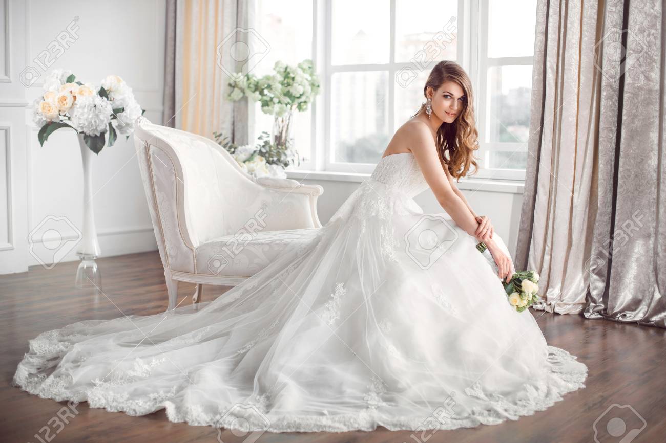 結婚式。自宅のような白いスタジオ インテリアの室内でソファーに座っている美しいドレスの花嫁。流行の結婚式のスタイルは、完全な長さで撮影。大きな窓に花嫁のような若い魅力的な白人ブルネット モデルは、ポーズを入札します。
