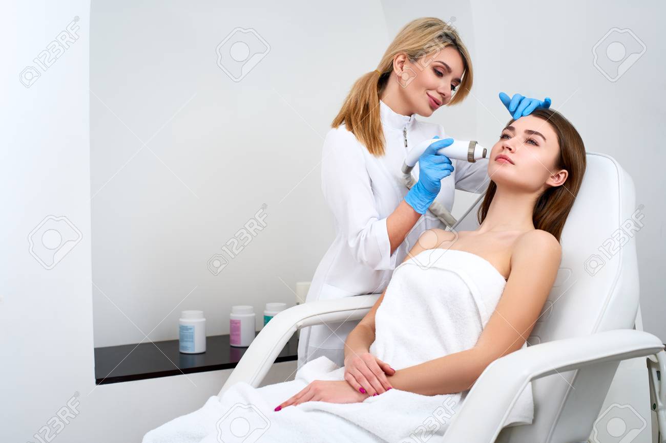 Pretty blonde beautician doctor doing rf-lifting procedure for woman laying in a beauty salon. Hardware cosmetology. Patient receiving electric facial massage. Skin rejuvenation and wrinkle smoothing. - 125263123 Pretty blonde beautician doctor doing rf-lifting procedure for woman laying in a beauty salon. Hardware cosmetology. Patient receiving electric facial massage. Skin rejuvenation and wrinkle smoothing. - 125263123