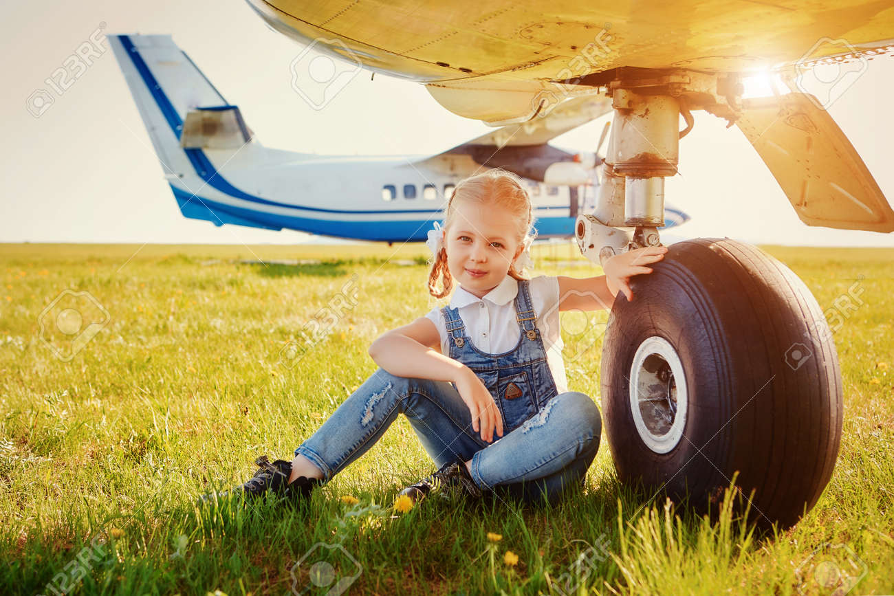 Cute little girl sitting near the landing gear wheels of an airplane and dreaming about traveling and flying in an airplane - 158561149 Cute little girl sitting near the landing gear wheels of an airplane and dreaming about traveling and flying in an airplane - 158561149