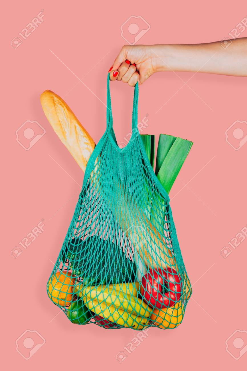 Woman hand holding a green mint string shopping bag with vegetables, fruits and bread in front of a pink background - 102747561 Woman hand holding a green mint string shopping bag with vegetables, fruits and bread in front of a pink background - 102747561
