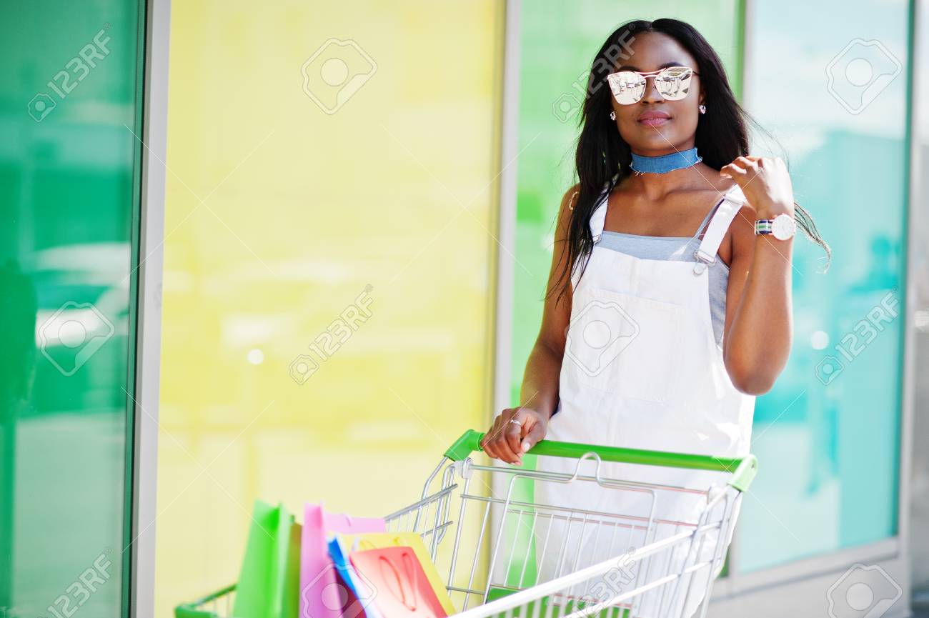 買い物カゴの横にある色とりどりの袋を保持しているサングラスと幻想的なアフリカ系アメリカ人女性のポートレート の写真素材 画像素材 Image