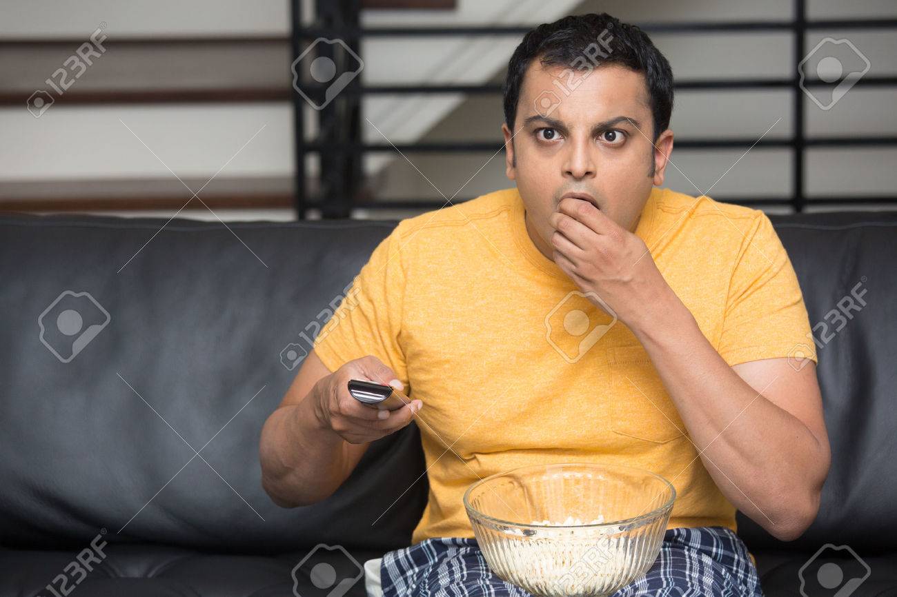 Closeup portrait, young man in yellow t-shirt, sitting on black leather couch, watching TV, holding remote, surprised at what he sees, munching popcorn, isolated indoors flat background - 47857647 Closeup portrait, young man in yellow t-shirt, sitting on black leather couch, watching TV, holding remote, surprised at what he sees, munching popcorn, isolated indoors flat background - 47857647