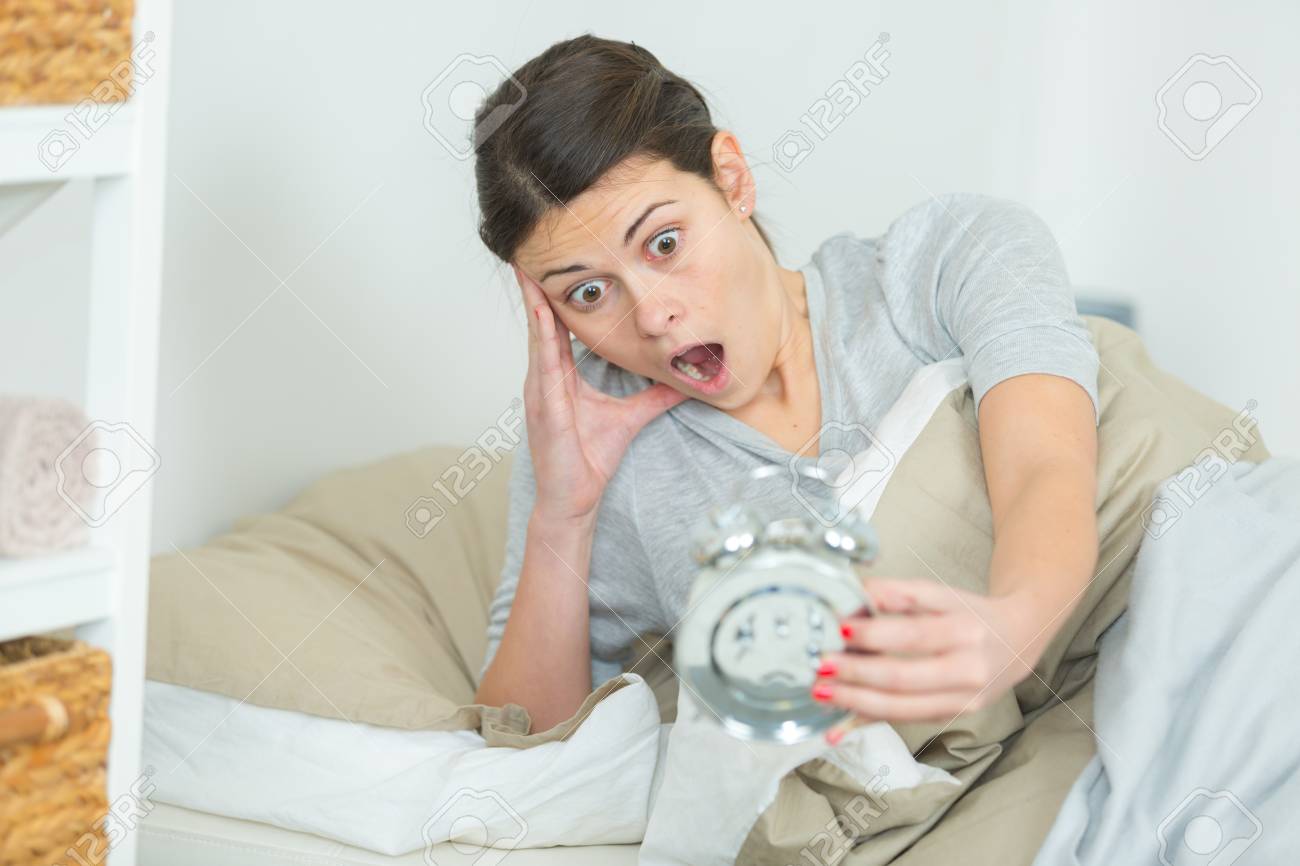 woman in bed looking at alarm clock with shocked expression stock