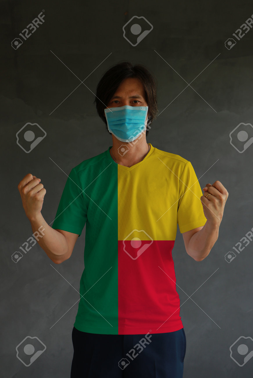 Man wearing hygienic mask and wearing Benin flag colored shirt and standing with raised both fist on dark wall background. Concept of protect tiny dust or disease. - 160156375 Man wearing hygienic mask and wearing Benin flag colored shirt and standing with raised both fist on dark wall background. Concept of protect tiny dust or disease. - 160156375