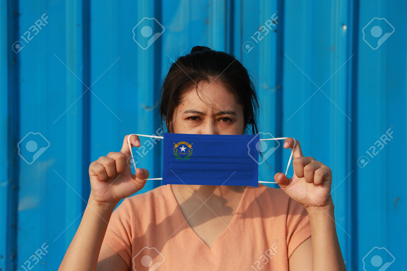 A woman and hygienic mask with Nevada flag pattern in her hand and raises it to cover her face on blue background. A mask is a very good protection from Tiny Particle or virus corona or Covid 19. - 160156378 A woman and hygienic mask with Nevada flag pattern in her hand and raises it to cover her face on blue background. A mask is a very good protection from Tiny Particle or virus corona or Covid 19. - 160156378