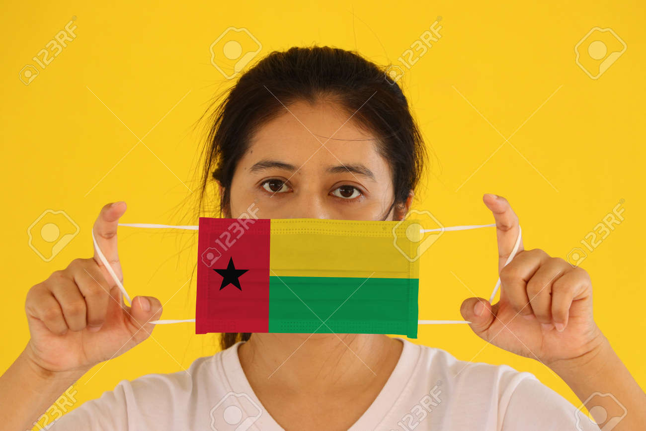 A woman in white shirt with Guinea bissau flag on hygienic mask in her hand and lifted up the front face on yellow background. - 165993609 A woman in white shirt with Guinea bissau flag on hygienic mask in her hand and lifted up the front face on yellow background. - 165993609