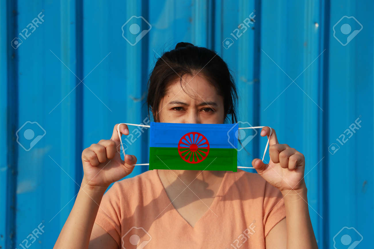 A woman with Romani people flag on hygienic mask in her hand and lifted up the front face on blue background. - 165993613 A woman with Romani people flag on hygienic mask in her hand and lifted up the front face on blue background. - 165993613