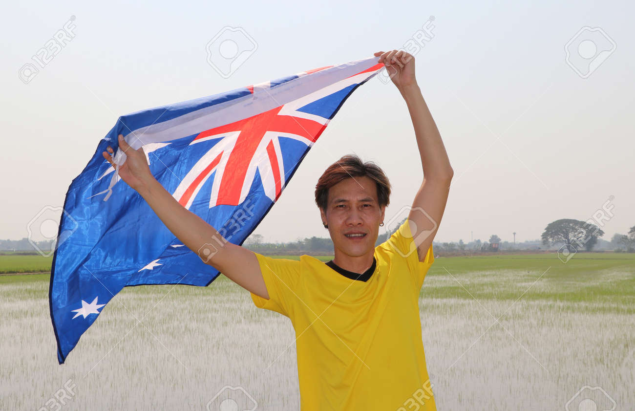 The man in yellow shirt is holding Australia flag in his hands and raising to the end of the arm at the back on nature view and rice fields background. - 163875069 The man in yellow shirt is holding Australia flag in his hands and raising to the end of the arm at the back on nature view and rice fields background. - 163875069