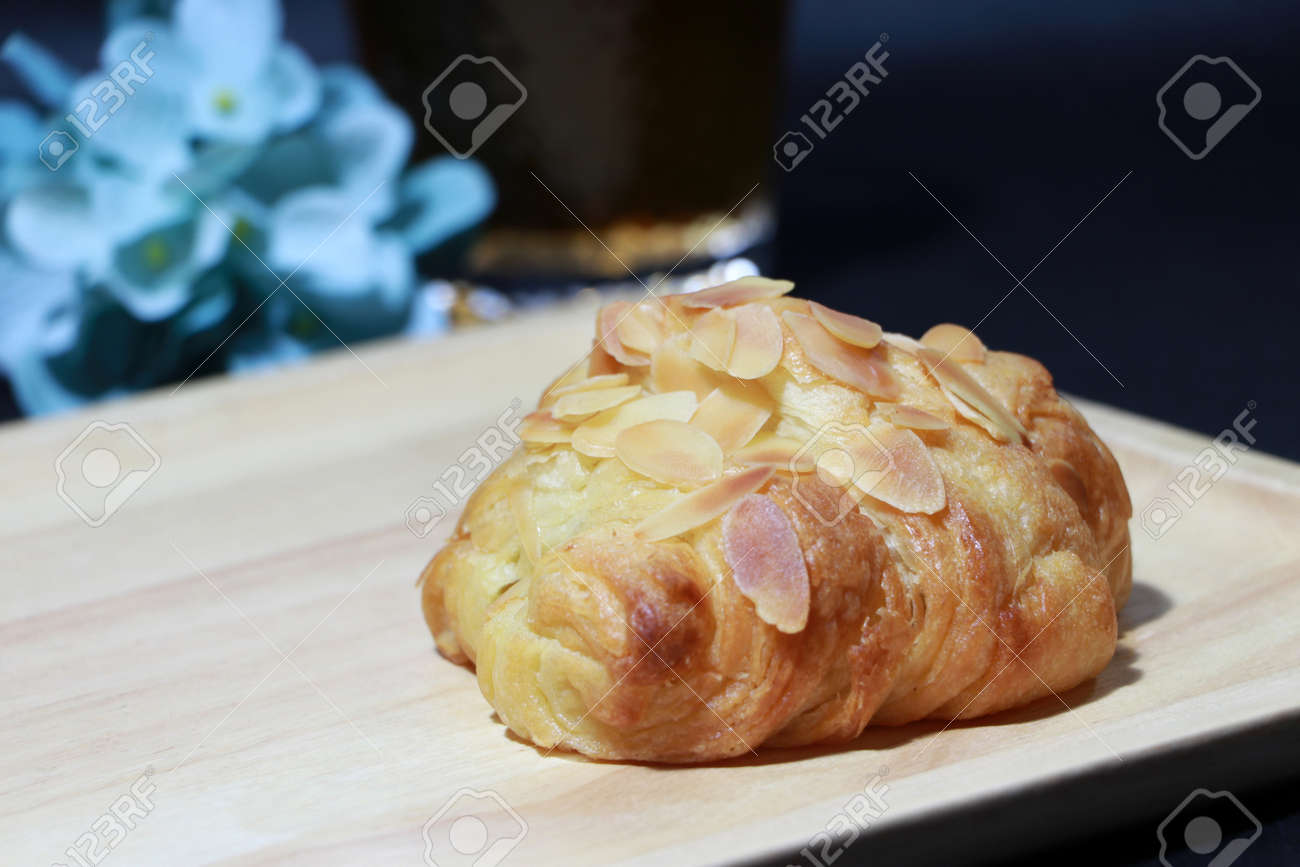 Almond flavor croissant placed on the square wooden tray with out focus tea in a glass and blue flowers on dark background. It is a French crescent shaped roll made of sweet flaky pastry, often eaten for breakfast. - 163505564 Almond flavor croissant placed on the square wooden tray with out focus tea in a glass and blue flowers on dark background. It is a French crescent shaped roll made of sweet flaky pastry, often eaten for breakfast. - 163505564