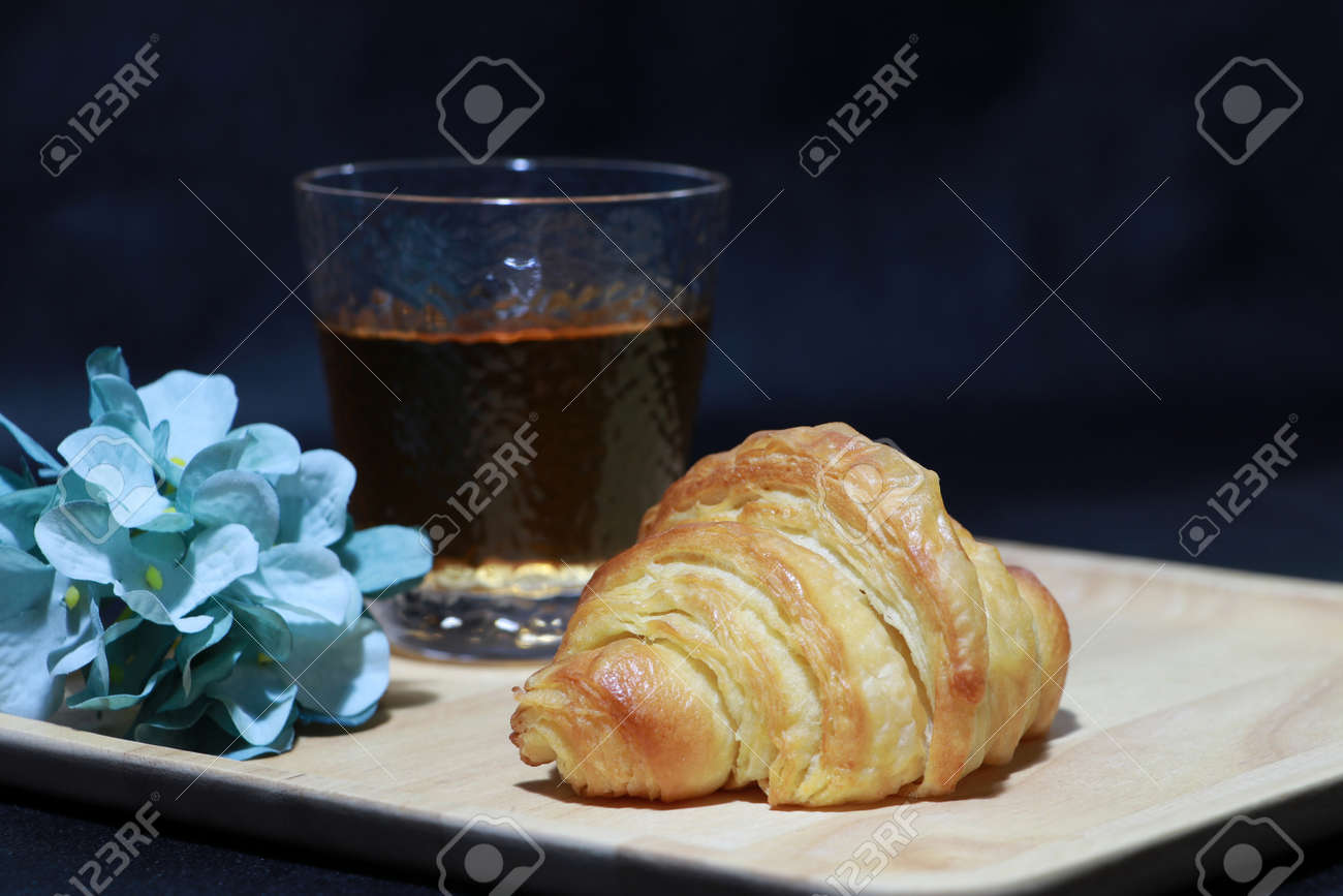 The croissant placed on the square wooden tray with tea in a glass and blue flowers on dark background. It is a French crescent shaped roll made of sweet flaky pastry, often eaten for breakfast. - 163505261 The croissant placed on the square wooden tray with tea in a glass and blue flowers on dark background. It is a French crescent shaped roll made of sweet flaky pastry, often eaten for breakfast. - 163505261