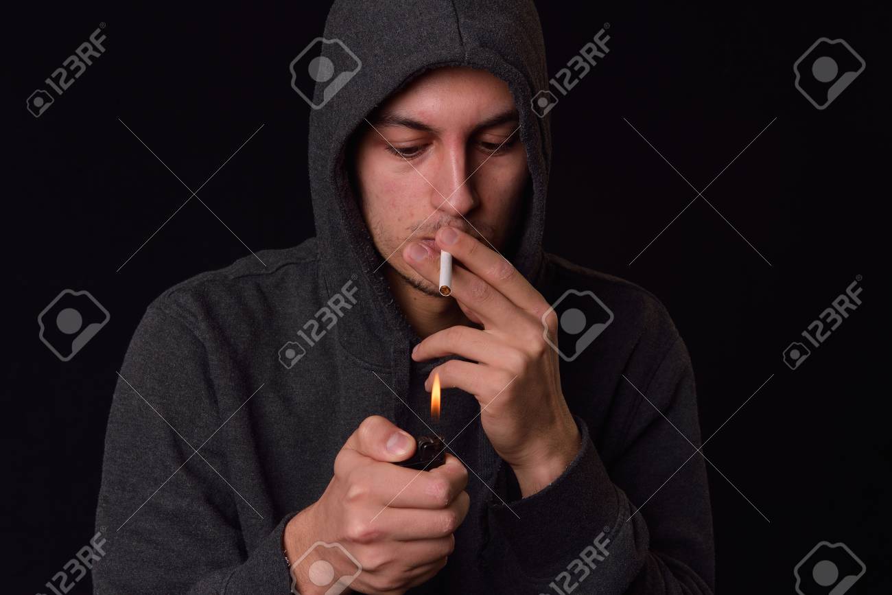 Teenager in a hooded sweatshirt lights a cigarette with lighter on a dark background. Harmful smoking concept. Unhealthy habits. Many others photos like this in my portofolio - 72765983 Teenager in a hooded sweatshirt lights a cigarette with lighter on a dark background. Harmful smoking concept. Unhealthy habits. Many others photos like this in my portofolio - 72765983