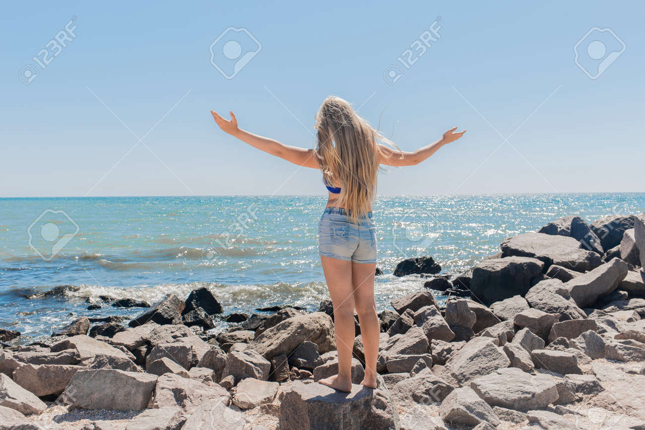 A Young Teenage Girl With Long Blonde Hair Blonde In Short Shorts Raised  And Spread Her Hands To The Sides Standing On The Rocks Near The Sea Shore  And The Beach. The