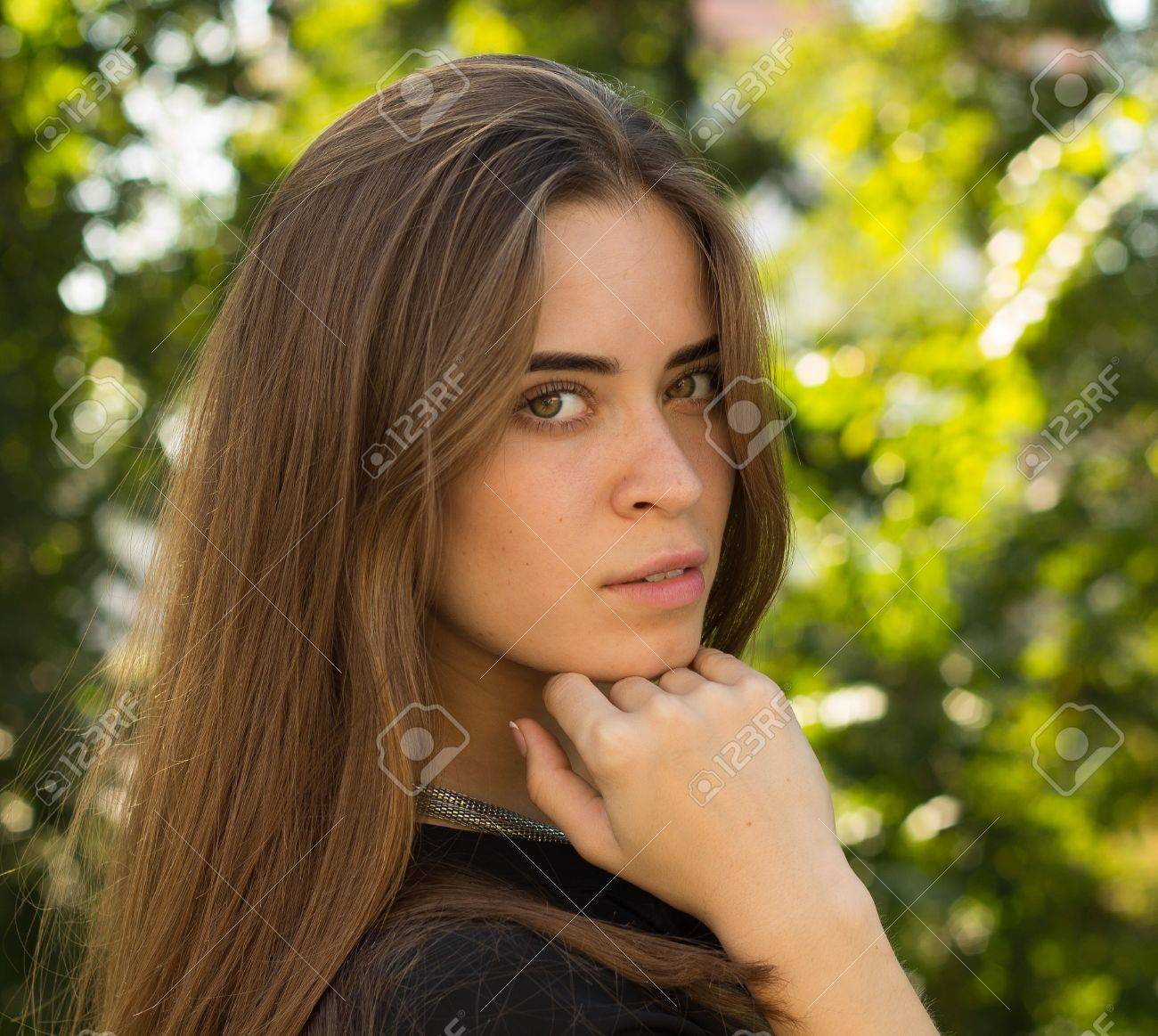 La Mujer Atractiva Joven Con El Largo De Color Marrón En La Blusa Negro Y Collar De De Pie Hacia Un En El Fondo De árboles Verdes Fotos, Retratos,