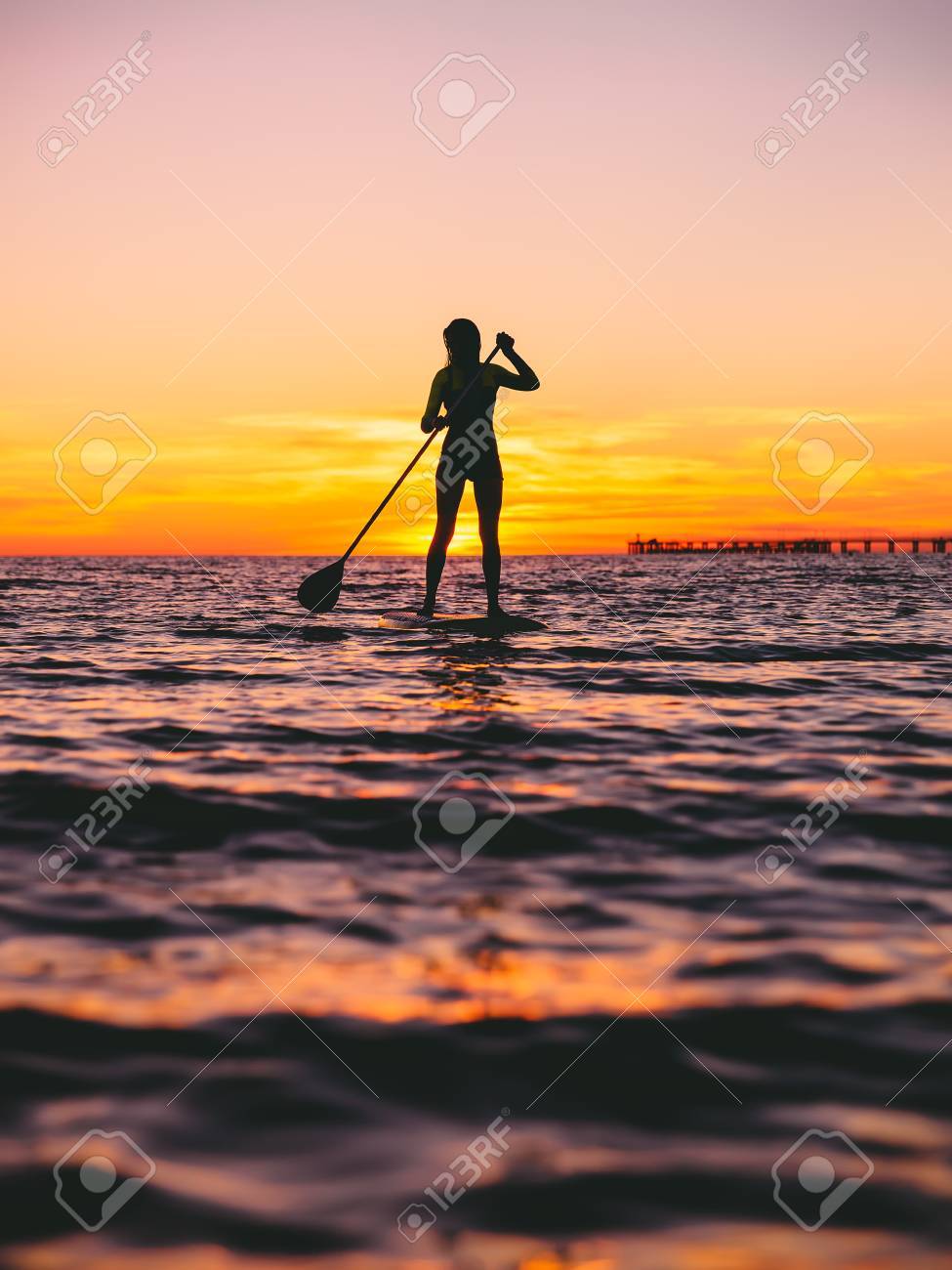 美しい夕日の色とフラット暖かい海で夕暮れ時に搭乗するパドルを立っている女性 の写真素材 画像素材 Image