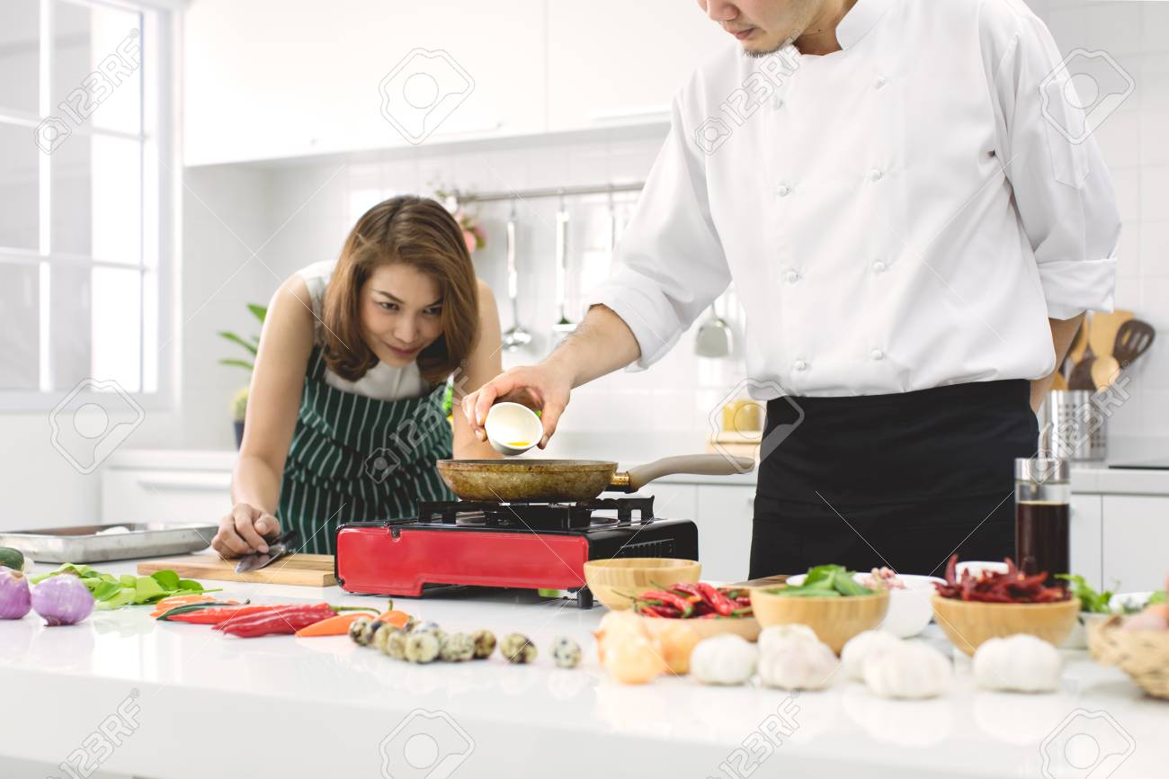 Beautiful Asian woman in red apron looking and noticing how to make fried egg, teaching by chef in white clean modern kitchen. - 112649619 Beautiful Asian woman in red apron looking and noticing how to make fried egg, teaching by chef in white clean modern kitchen. - 112649619