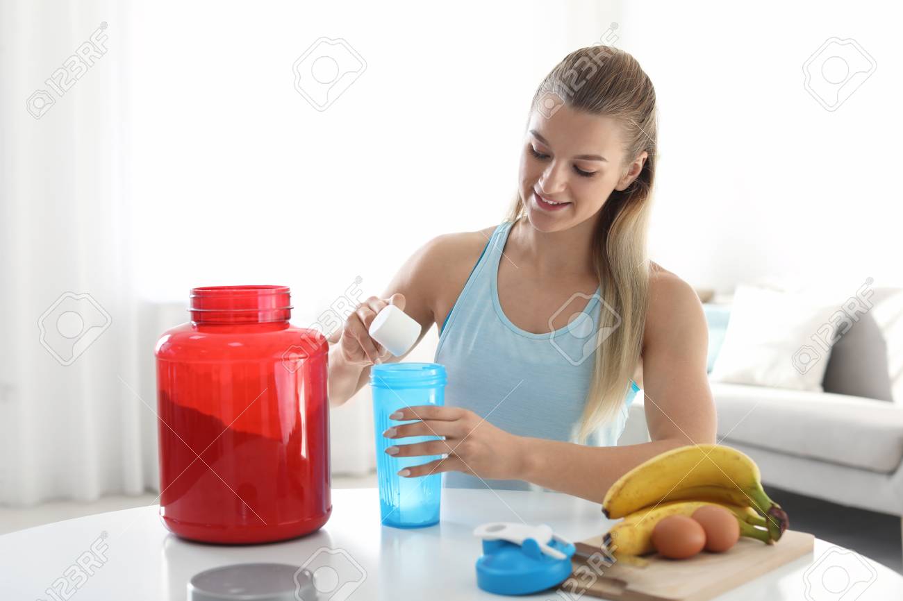 Young woman preparing protein shake at table in room - 114844635 Young woman preparing protein shake at table in room - 114844635