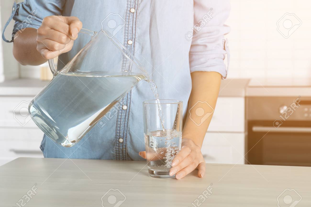 Woman pouring water into glass in kitchen, closeup. Refreshing drink - 117117981 Woman pouring water into glass in kitchen, closeup. Refreshing drink - 117117981