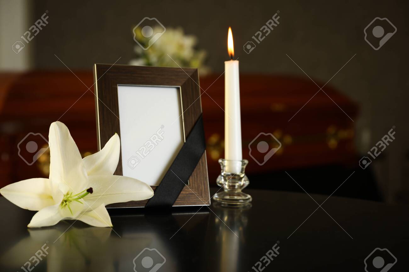 Black photo frame with burning candle and white lily on table in funeral home - 128458331 Black photo frame with burning candle and white lily on table in funeral home - 128458331